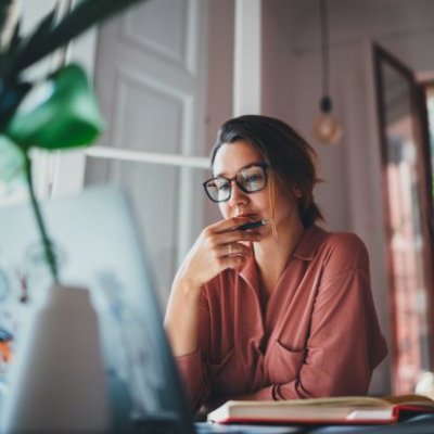 A woman in glasses sits at a desk in a home office looking worried.